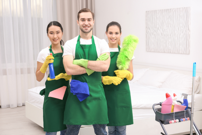 Three people in aprons smiling while holding various cleaning supplies, ready to tackle a cleaning task together.